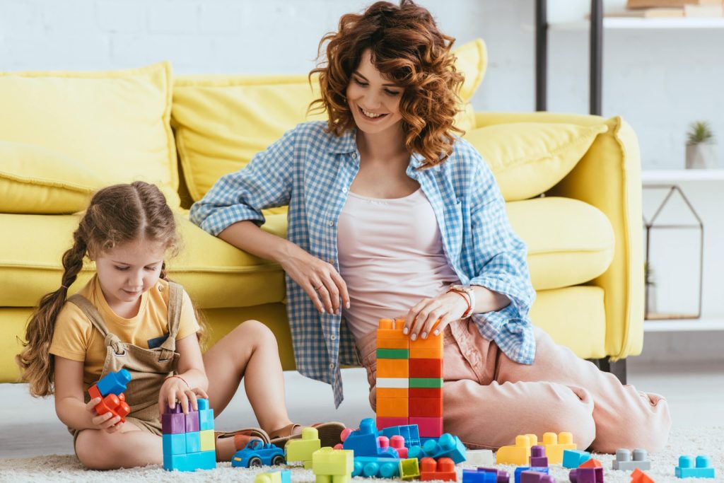 Une petit fille assise sur un tapis devant un canapé joue aux Lego, accompagnée par sa nounou qui la regarde faire en souriant